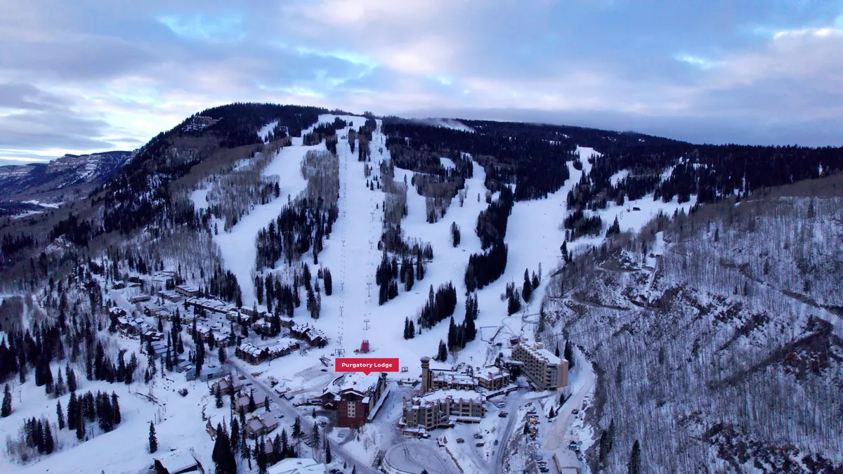 A striking exterior perspective of Purgatory Lodge with snow-covered rooftops, surrounded by dramatic mountains and tree-lined ski slopes. The lodge stands in the heart of the resort, with the slopes just steps away, evoking the excitement of ski-in/ski-out convenience. The snowy landscape and warm glow from lodge windows set a cozy, adventurous mood.
