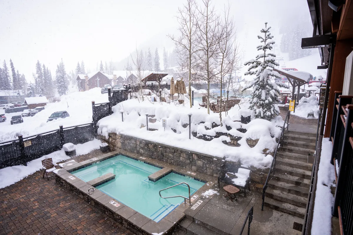 An inviting exterior shot of a steaming hot tub nestled among snow-covered trees, with a stone wall and steps leading down for access. The tranquil setting is perfect for unwinding after a day on the slopes, echoing the spa-like and adult swim spa amenities.