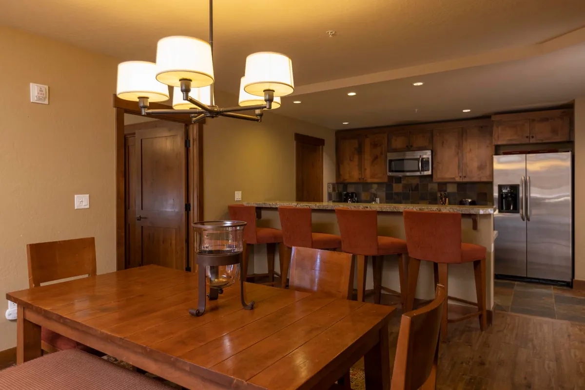 A spacious, open-concept dining area with a granite-topped table, elegant chandelier, and adjacent kitchen counter with bar stools. The setting is styled for both intimate family meals and lively gatherings, with plenty of natural light and a warm, welcoming mood.