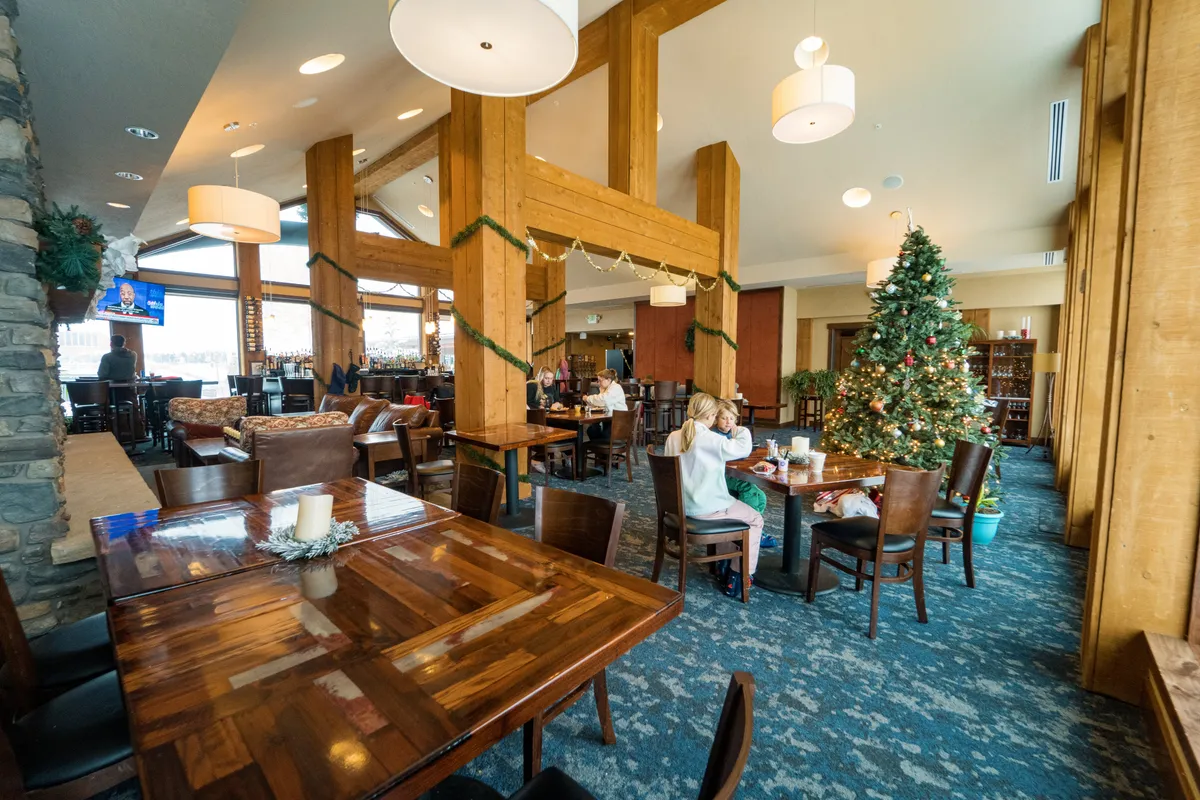 A festive great room with large wooden beams, a decorative Christmas tree, dining tables set for gatherings, and a cozy bar area. The space exudes warmth and togetherness, ideal for family celebrations and casual get-togethers.