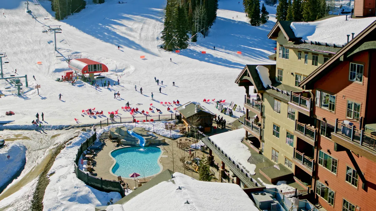 A lively exterior scene showing both the swimming pool and adjacent ski slopes, with guests visible swimming, skiing, and relaxing by the lodge. Snow blankets the landscape, and the pool area stands out as a focal point for activity and relaxation.