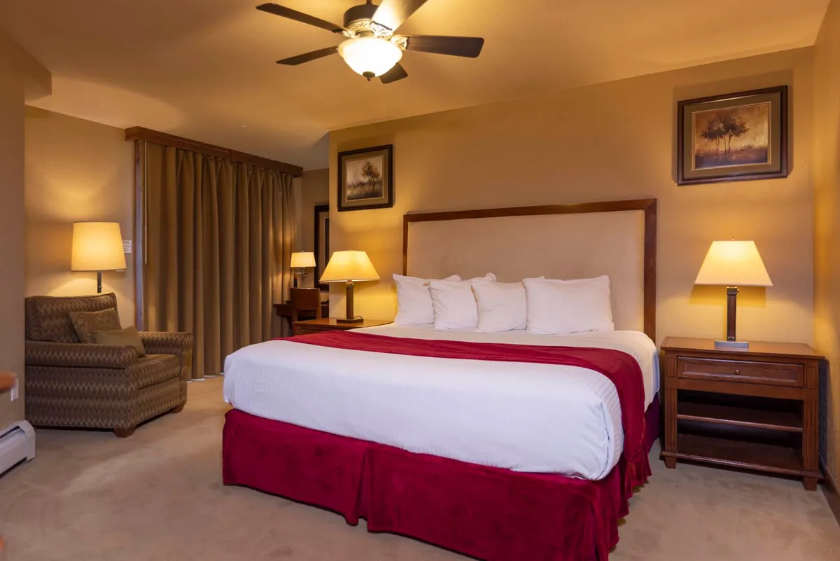 This image features a spacious king-sized bed adorned with plush linens and red accents, flanked by wooden nightstands with softly glowing lamps. A cozy armchair sits invitingly by the window, basking in natural morning light, creating an inviting nook for reading or unwinding. The overall atmosphere exudes restful luxury and warmth.