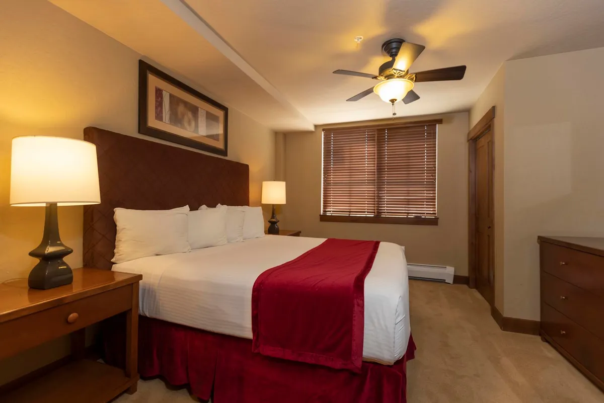 The cozy bedroom features a bed with a bold red accent, a softly lit nightstand, and a wooden dresser. A window offers glimpses of the outdoors, suggesting a peaceful place to relax after a day of adventure. The ambiance is intimate and inviting, perfect for couples.