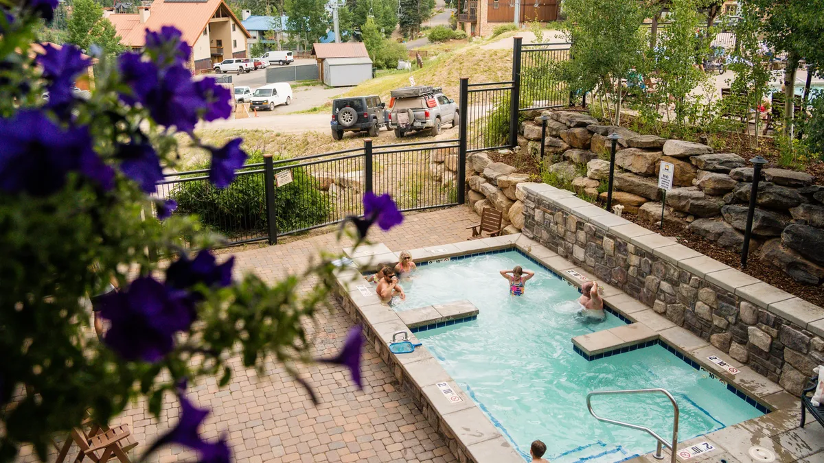 An inviting outdoor pool area with a hot tub, surrounded by natural stonework, blooming flowers, and guests enjoying a swim and relaxation. The scene suggests family fun and post-ski relaxation options, with the pool and hot tub as highlights.